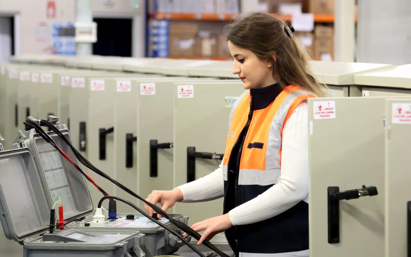 A technician in a high-visibility vest works with test equipment beside a row of electrical cabinets.